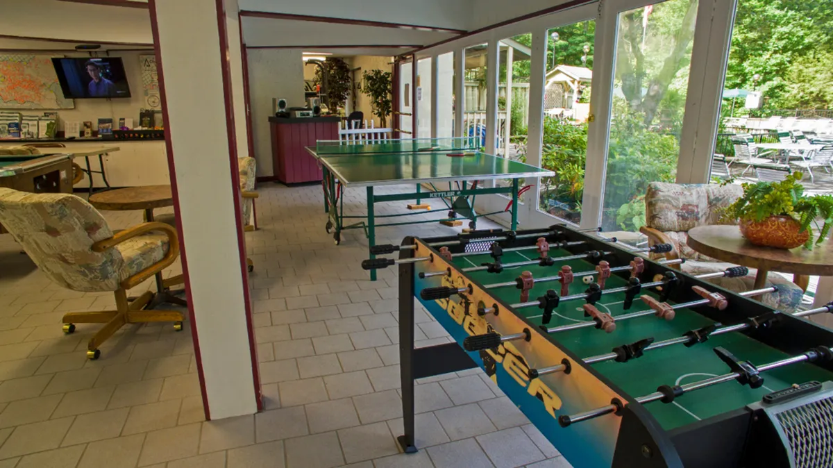 A lively and casual game room interior featuring a foosball table, ping pong table, television, and a comfortable seating area. The space is designed for friendly competition, group play, and casual gatherings.