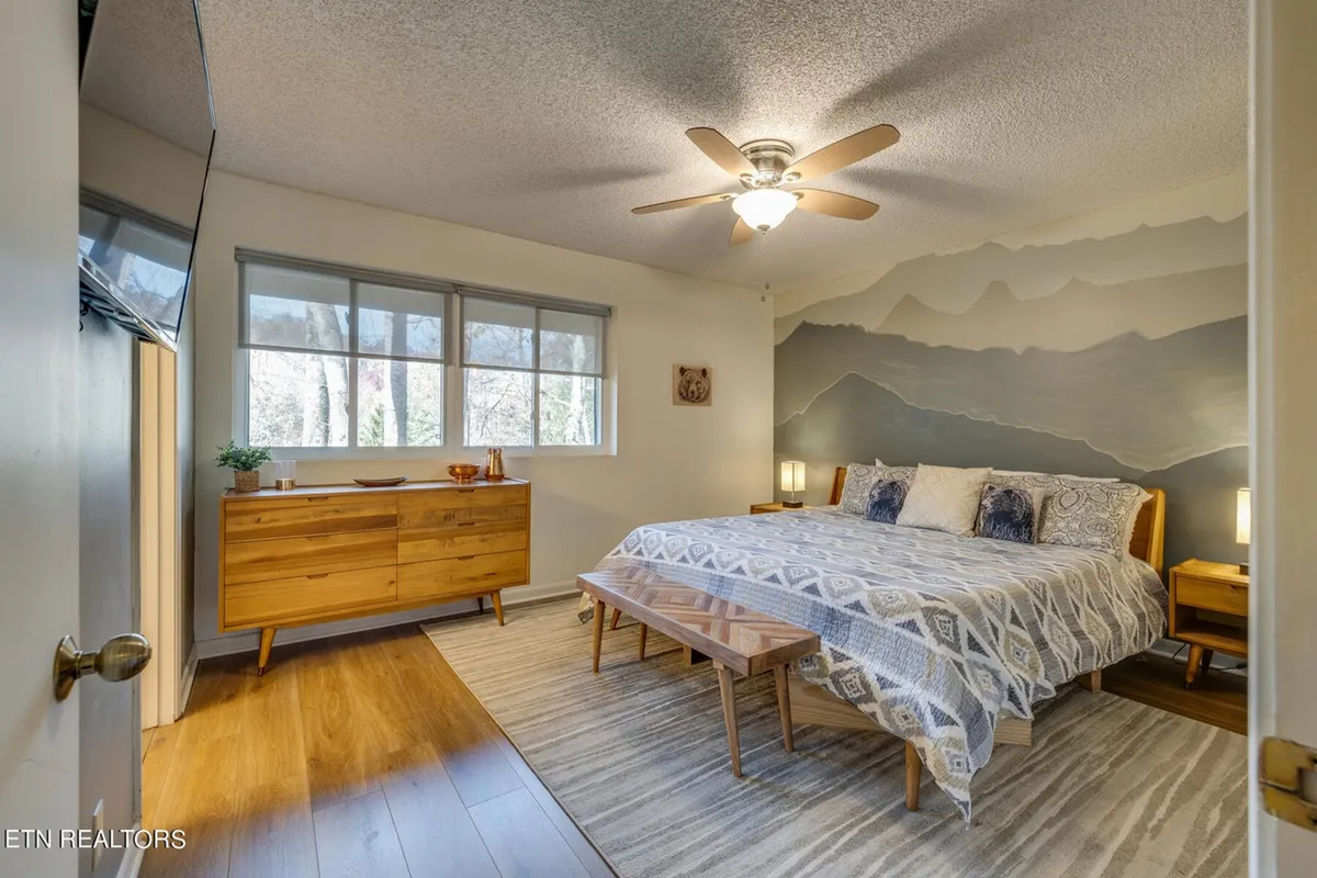 A serene bedroom featuring a large bed with plush bedding, flanked by nightstands and a dresser. A tranquil mountain mural sets a soothing ambiance, and soft lighting enhances the sense of relaxation. Natural light filters in, contributing to the room’s peaceful atmosphere.