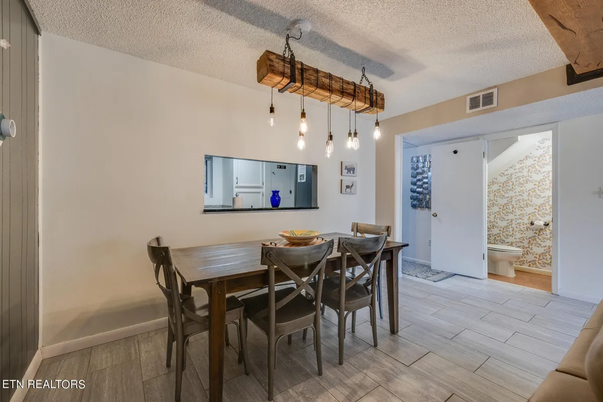 A cozy dining area centered around a sturdy dining table, adorned with a striking hanging light fixture. The setup is warm and communal, with a decorative mirror and a blue vase adding stylish touches.