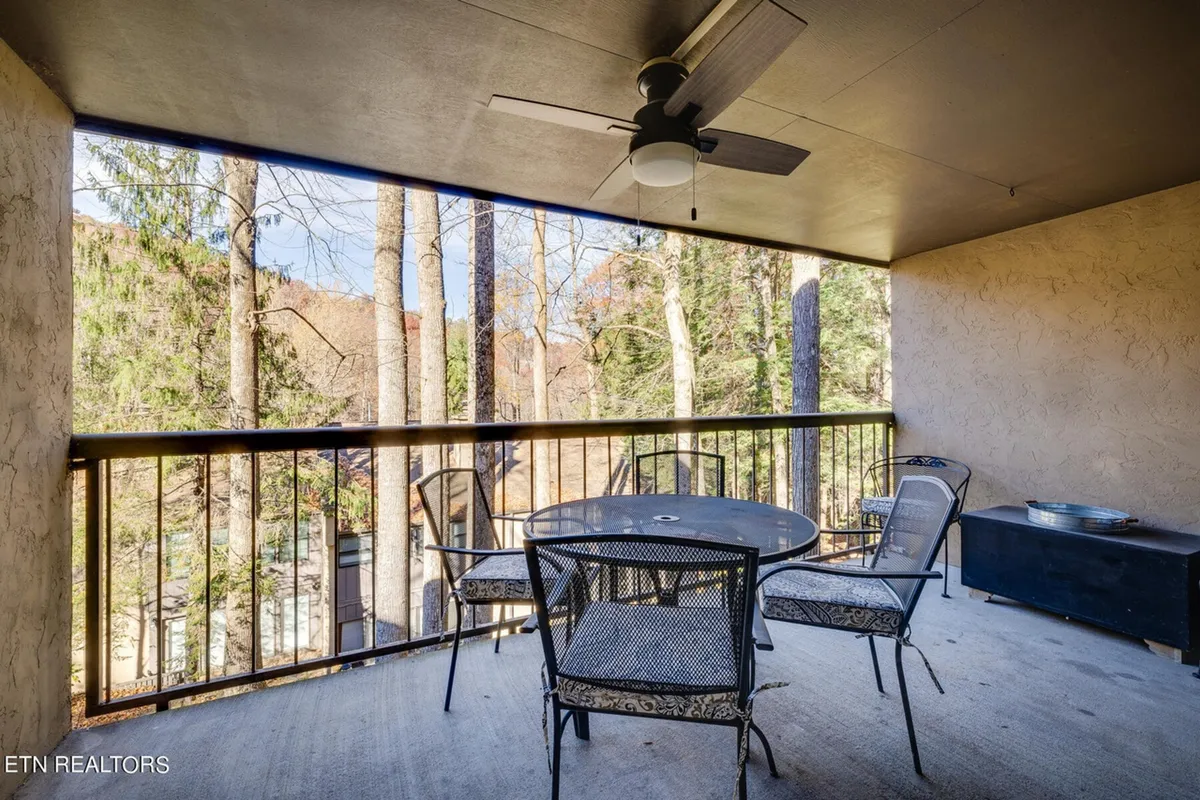 A cozy, tranquil outdoor seating area with a table and comfortable chairs, set on a patio or balcony. Trees provide a scenic, private backdrop, and a ceiling fan hangs above, reinforcing comfort and relaxation. The setting is ideal for enjoying morning coffee, evening conversations, or simply unwinding while soaking in peaceful views.