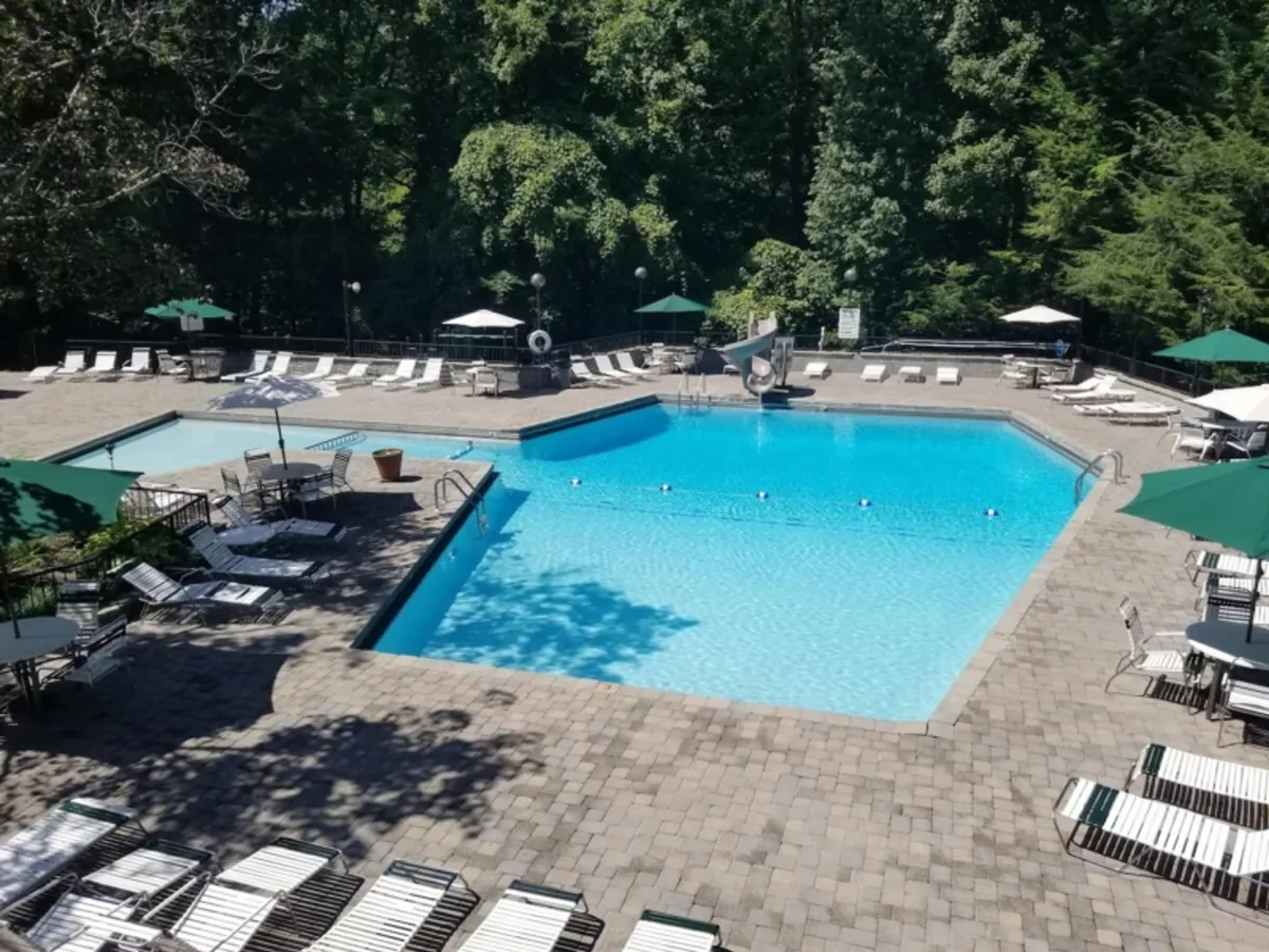 A cheerful outdoor resort pool scene featuring a water slide, lounge chairs, and umbrellas surrounded by lush greenery. Children and families are seen enjoying the pool, emphasizing play and relaxation in a bright, inviting setting.