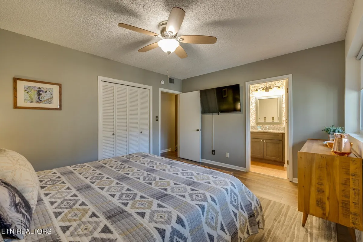 A bright and comfortable bedroom showcasing a bed with patterned bedding, a wooden dresser, wall-mounted TV, and framed art. The space is bathed in natural light, offering a modern yet welcoming feel for relaxation and leisure.