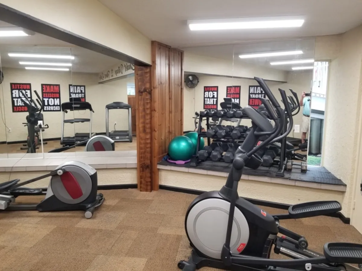 A well-equipped gym interior includes an elliptical machine, treadmill, free weights, an exercise ball, and mirrored walls. The space is designed for both solo and group workouts, exuding a clean and motivating atmosphere.