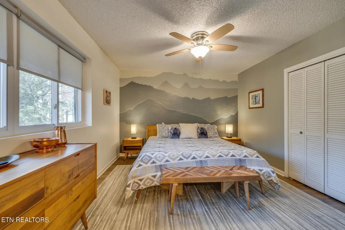 A cozy bedroom with a bed adorned in patterned bedding, complemented by wooden furniture. The space features a mountain mural, soft natural lighting, and homey accents, creating an inviting environment ideal for couples or families.