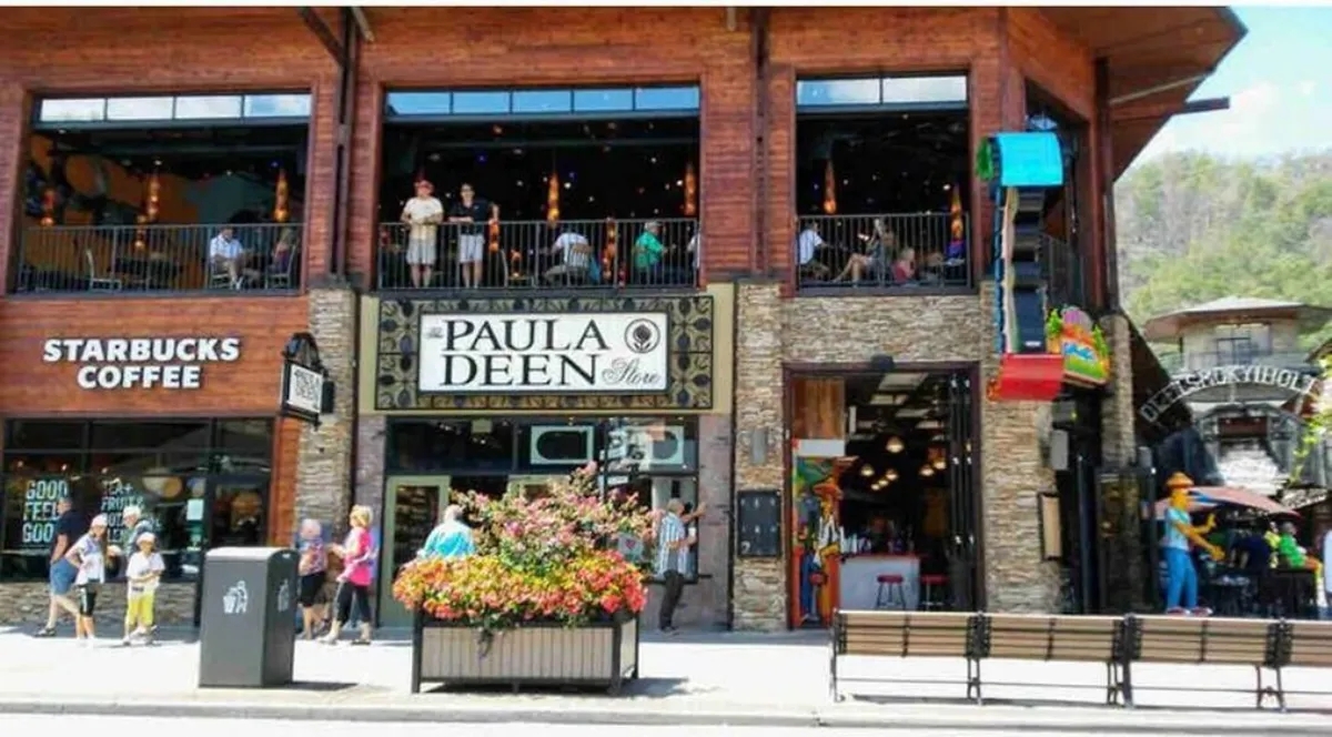 A vibrant streetscape showing the Paula Deen Store and Starbucks Coffee signage, flower arrangements, and people dining or strolling. The atmosphere is energetic and inviting, reflecting the bustling vibe of downtown Gatlinburg.