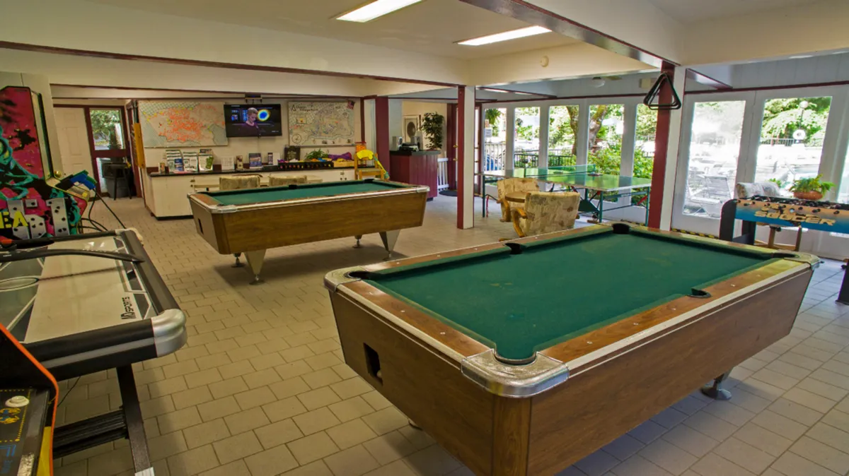 An inviting interior view of the game room at the Owners Club, featuring pool tables, an air hockey table, a TV, and comfortable seating. The setting is bright and energetic, with people engaging in friendly games and socializing in a laid-back environment.