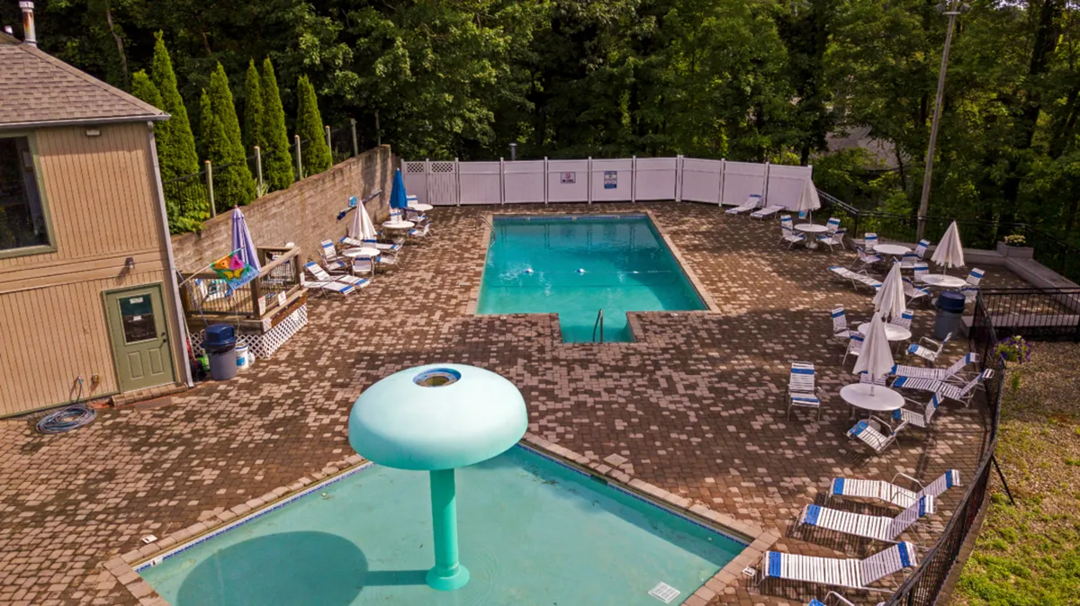 An inviting outdoor swimming pool area surrounded by trees, with a water feature, lounge chairs, and children playing in the water. The sun is setting, casting a warm glow over the scene, and the setting encourages socializing, swimming together, and relaxing in a resort-like environment.