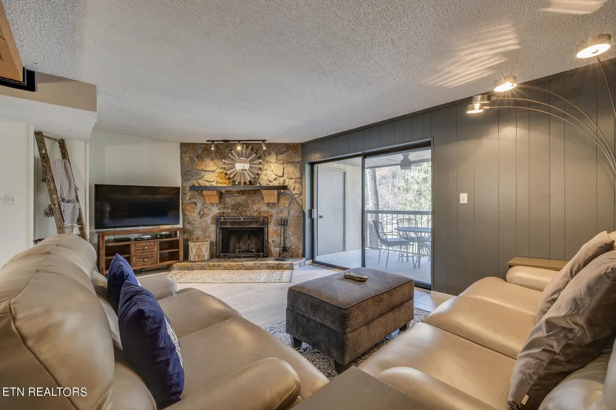 A bright, open-concept living space with a stone fireplace, decorative rug, comfortable seating, and generous windows that allow natural light to pour in and provide beautiful mountain or forest views.