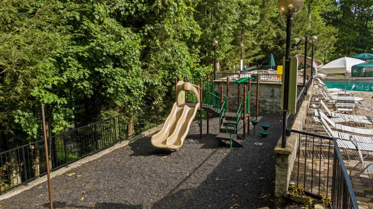 A vibrant exterior shot capturing the Chalet Village Owners Club pool area with children playing on the playground equipment nearby. The scene is filled with lush greenery, a sparkling swimming pool, and relaxed seating areas where families gather and socialize. The overall atmosphere highlights active outdoor fun and family togetherness.