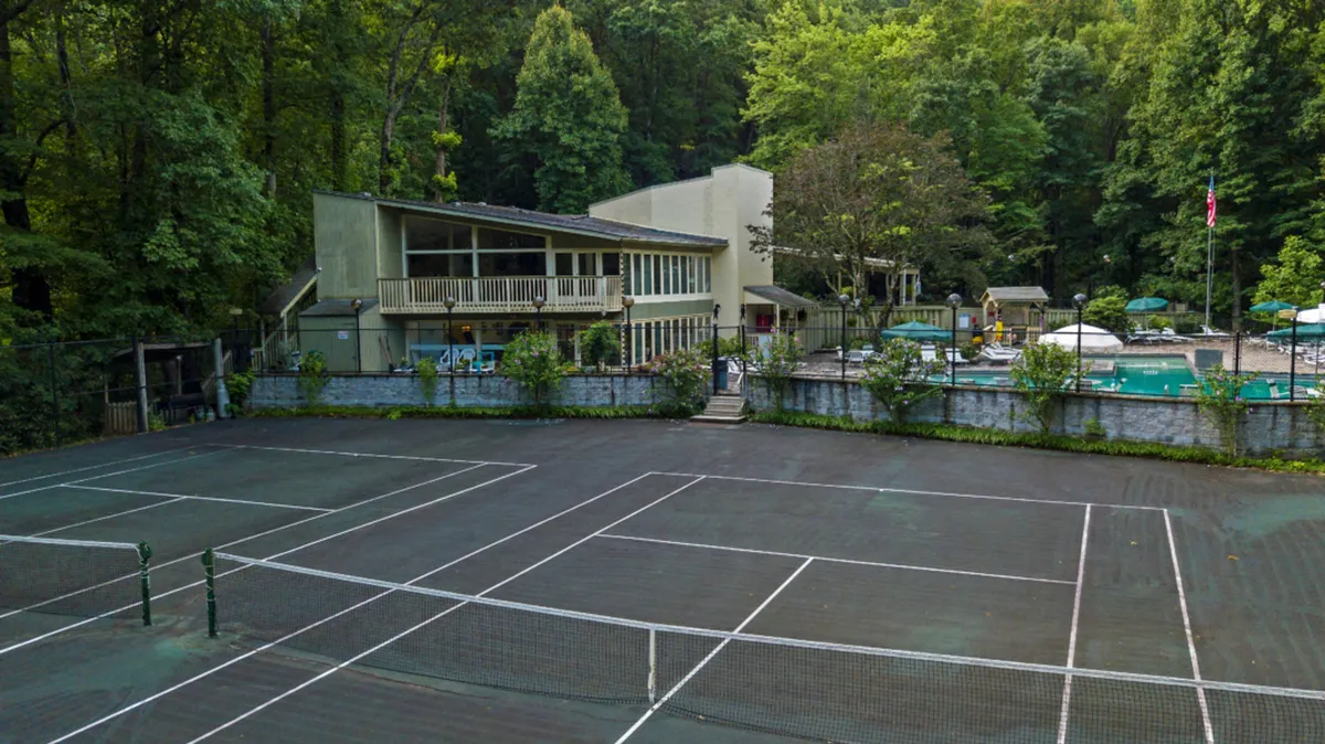 A vibrant exterior view of the resort complex, highlighting tennis courts, a sparkling outdoor swimming pool, and the main building bustling with activity. Guests are seen playing tennis, swimming, and relaxing outdoors, suggesting a lively, communal atmosphere.
